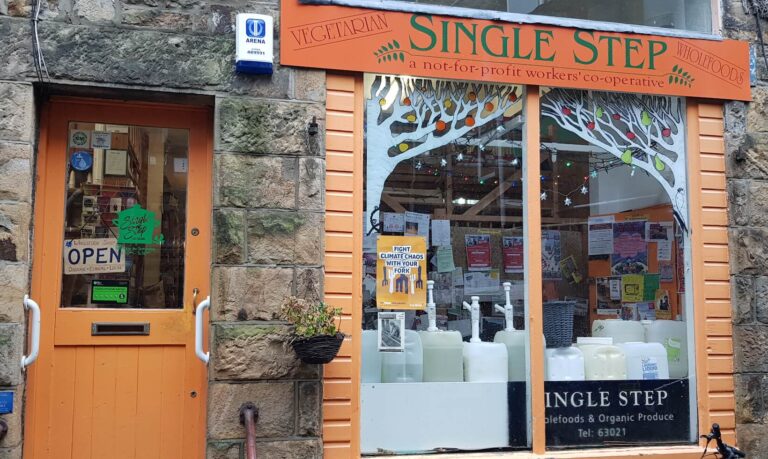 Front entrance of Single Step, a vegetarian wholefoods shop and not-for-profit workers' co-operative in a stone building, featuring an orange-framed window with painted fruit tree, posters inside, and refillable containers visible through the glass.